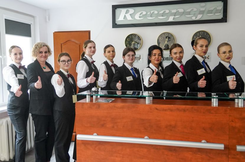 Na zdjęciu widać uczniów wraz z nauczycielami z klasy hotelarskiej przy ladzie w recepcji kompleksu hotelowego