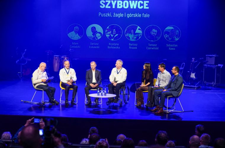 Rozwiń skrzydła z Fundacją PODAJ DALEJ. Na zdjęciu, w niebieskim oświetleniu, widać uczestników rozmowy siedzących na krzesłach w półkolu na scenie Oskardu.