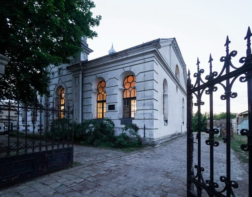 Architektoniczna perełka odzyska blask. Na zdjęciu zrobionym o zmierzchu widać otwartą bramę wjazdową, za nią w tle jest synagoga z rozświetlonymi oknami. Z lewej strony fragment budynku zasłania drzewo.