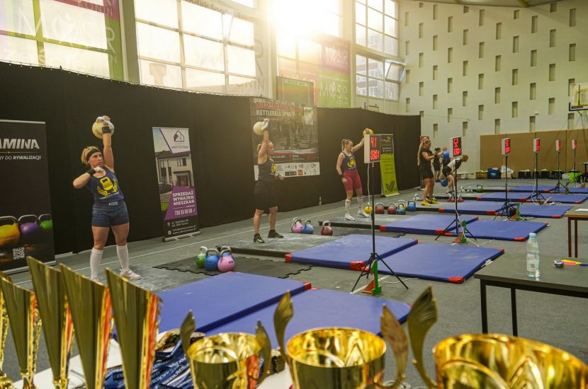 Zdjęcie przestawia puchary oraz rywalizujące zawodniczki podczas Mistrzostw Polski Kettlebell