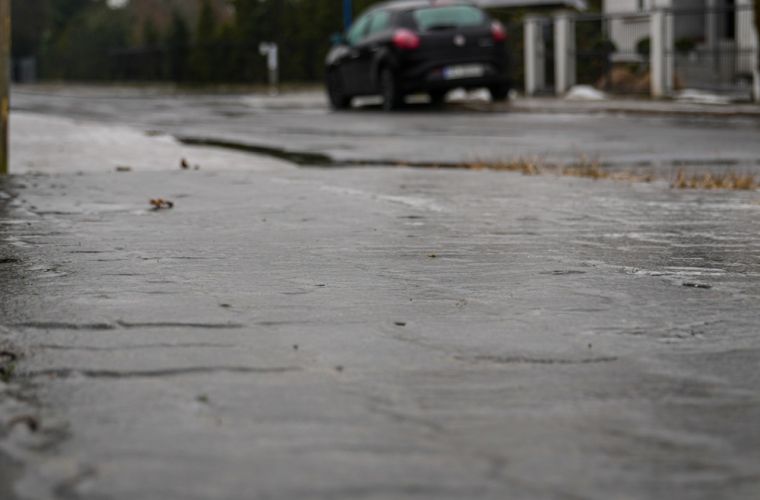Uwaga, marznące opady deszczu! Na zdjęciu wykonanym z perspektywy żaby widać śliską nawierzchnię ulicy i stojący w oddali, na poboczu, samochód.
