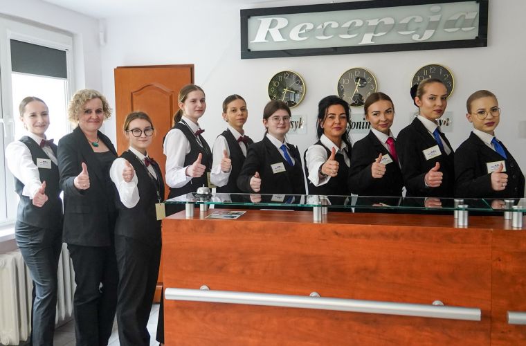Na zdjęciu widać uczniów wraz z nauczycielami z klasy hotelarskiej przy ladzie w recepcji kompleksu hotelowego
