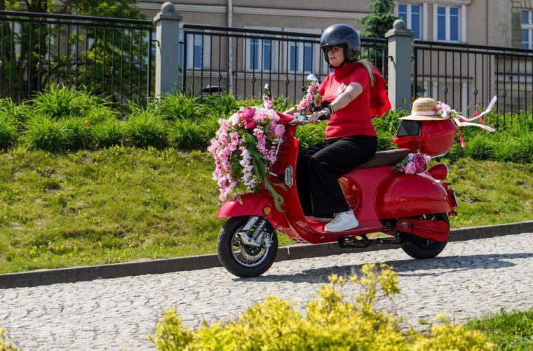 Zdjęcie przedstawia panią jadącą na różowym skuterze przyozdobionym kwiatami