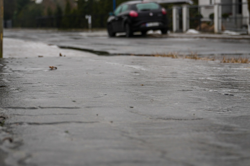 Uwaga, marznące opady deszczu! Na zdjęciu wykonanym z perspektywy żaby widać śliską nawierzchnię ulicy i stojący w oddali, na poboczu, samochód.
