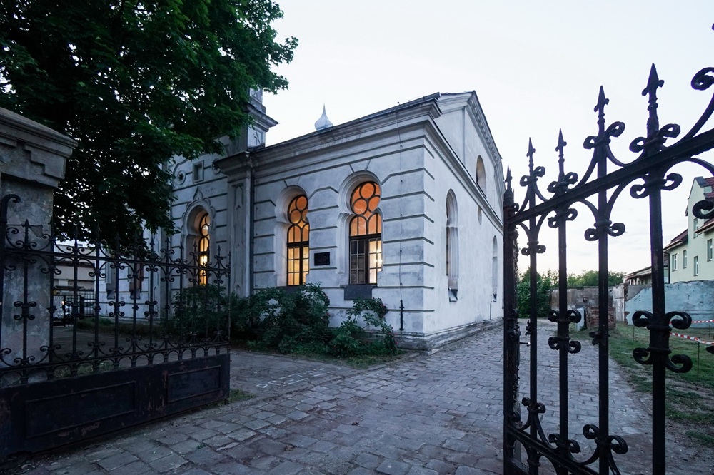Architektoniczna perełka odzyska blask. Na zdjęciu zrobionym o zmierzchu widać otwartą bramę wjazdową, za nią w tle jest synagoga z rozświetlonymi oknami. Z lewej strony fragment budynku zasłania drzewo.