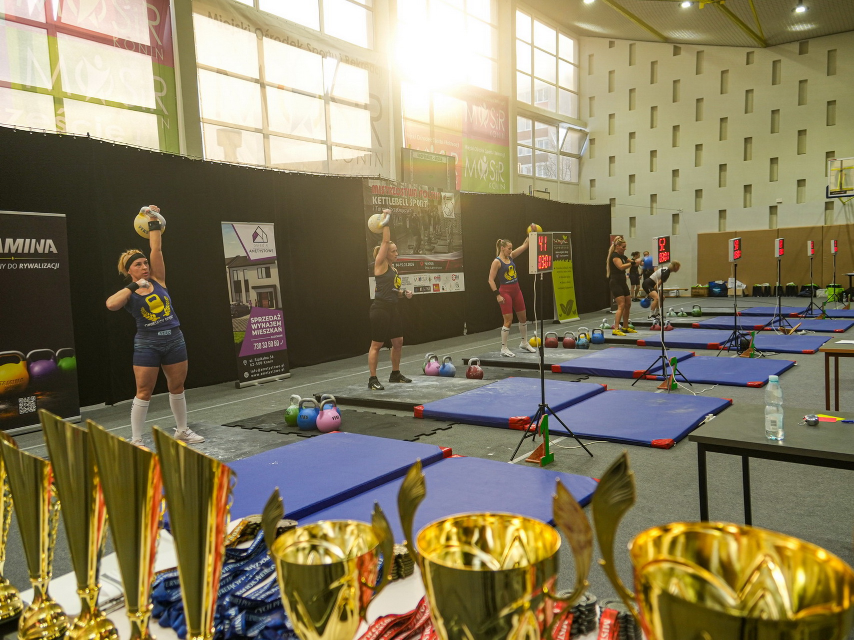 Zdjęcie przestawia puchary oraz rywalizujące zawodniczki podczas Mistrzostw Polski Kettlebell