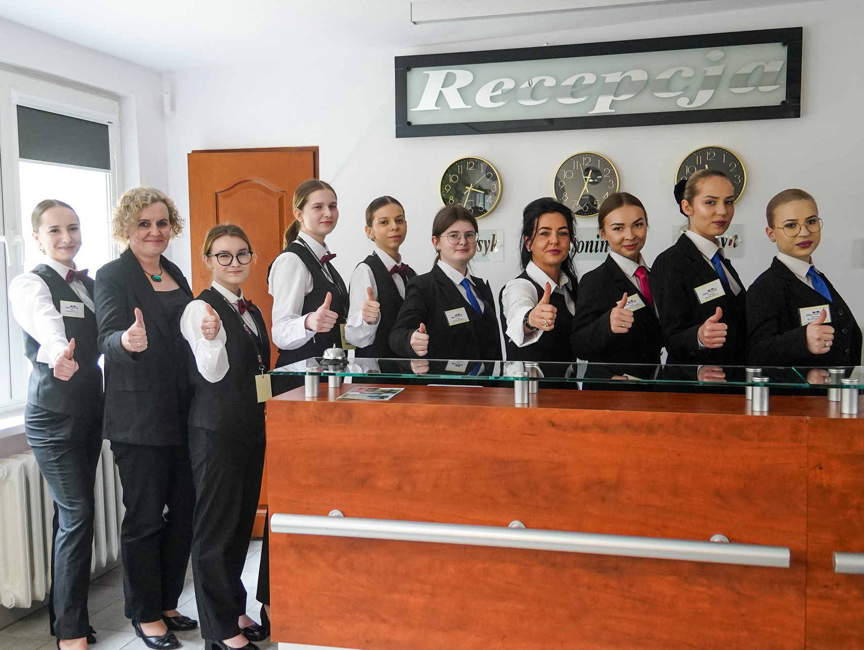Na zdjęciu widać uczniów wraz z nauczycielami z klasy hotelarskiej przy ladzie w recepcji kompleksu hotelowego