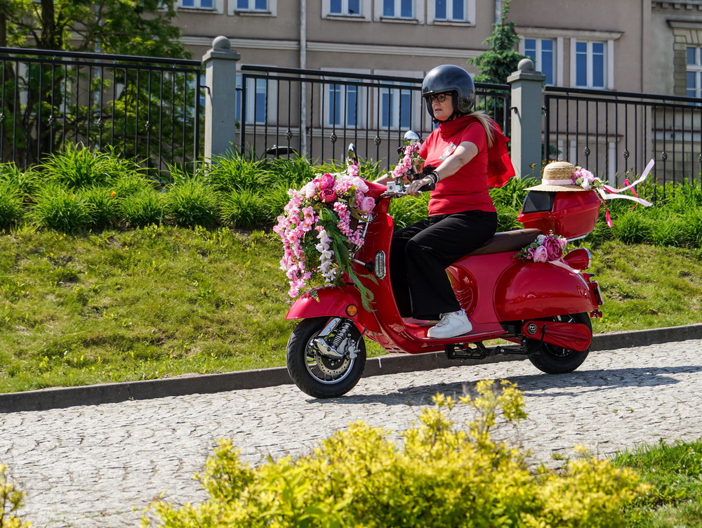Zdjęcie przedstawia panią jadącą na różowym skuterze przyozdobionym kwiatami